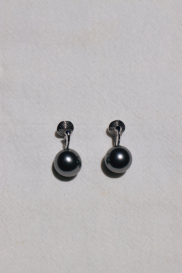 An overhead shot of a pair of dark gray spherical pearl drop earrings, each with a unique spiral-shaped silver-toned top. They are resting on a white textured fabric.