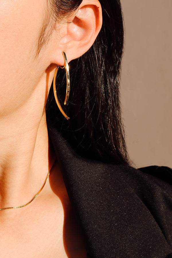 A close-up side profile of a woman's ear, showcasing a shiny gold curved hoop earring. The woman has long dark hair and is wearing a black blazer. A thin gold chain necklace is visible around her neck, with sunlight casting shadows.