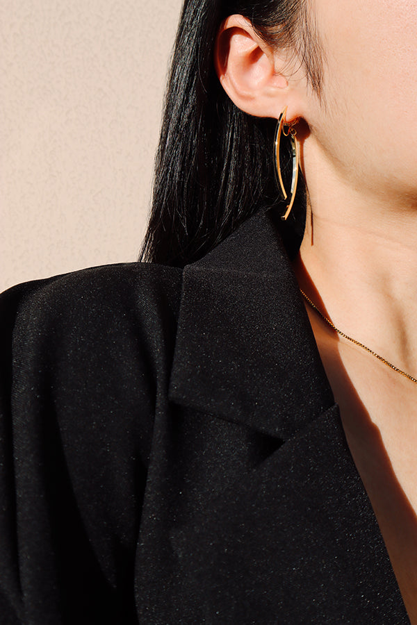 A close-up side profile of a woman's ear and shoulder, showcasing a shiny gold curved hoop earring. The woman has long dark hair and is wearing a black blazer. A thin gold chain necklace is visible around her neck, with sunlight creating a bright gleam on the earring.