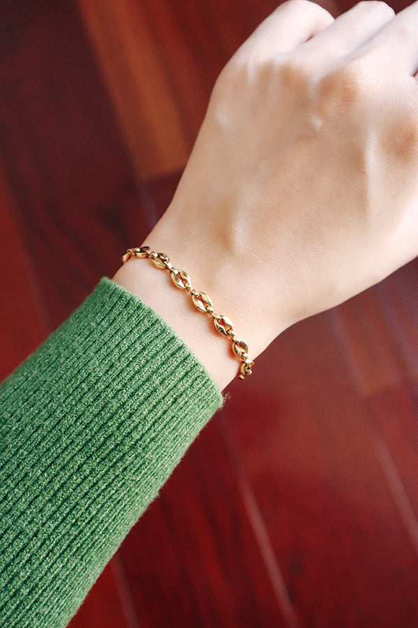 A close-up of a woman's wrist, wearing a gold-toned link bracelet with a coffee-bean design. The gold tone is set against a green ribbed sweater sleeve and a blurred reddish-brown wooden floor background.