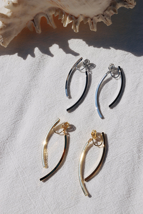 An overhead shot of two pairs of curved bar earrings on a white textured fabric, with a large seashell in the background. The top pair is silver-toned, and the bottom pair is gold-toned, both designed to hook onto the ear.