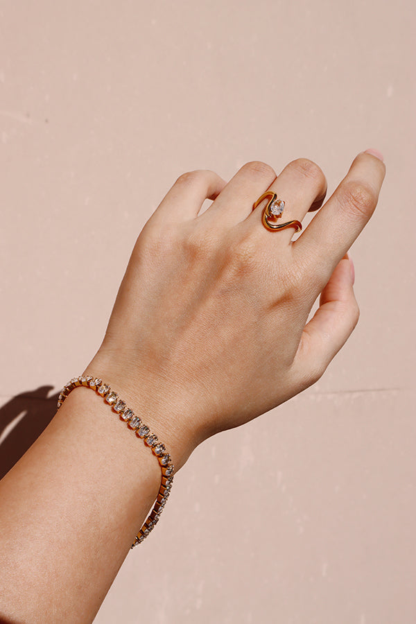 A woman's arm and hand, wearing the Eveline Curve Ring on her ring finger and a dazzling gold tennis bracelet encrusted with clear stones on her wrist. The background is a flat, light pink surface.