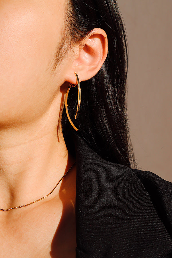 A close-up side profile of a woman's ear, showcasing a shiny gold curved hoop earring. The woman has long dark hair and is wearing a black blazer. A thin gold chain necklace is visible around her neck. Sunlight casts a strong highlight on the earring.