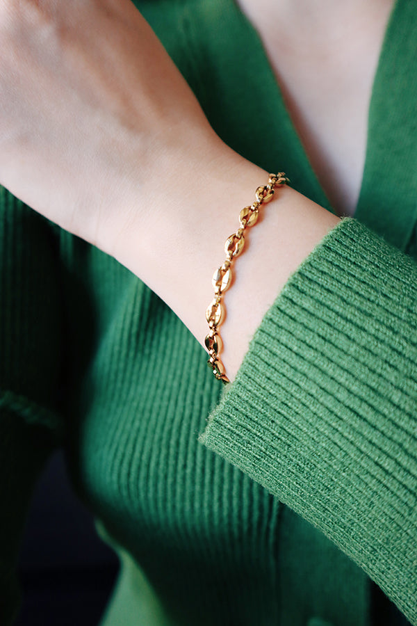 A close-up of a woman's wrist, wearing the gold-toned Isla Everyday Link Bracelet. The coffee-bean link design contrasts nicely against the woman's vibrant green ribbed sweater.