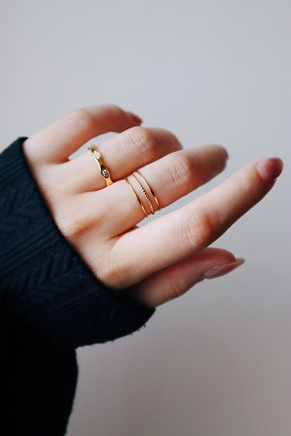 A close-up of a woman's hand showcasing a ring stack. The index finger wears the Amara Sparkle Ring (wavy band with stones), and the middle finger wears three layered gold bands, two smooth and one beaded. The woman is wearing a black long-sleeved top.