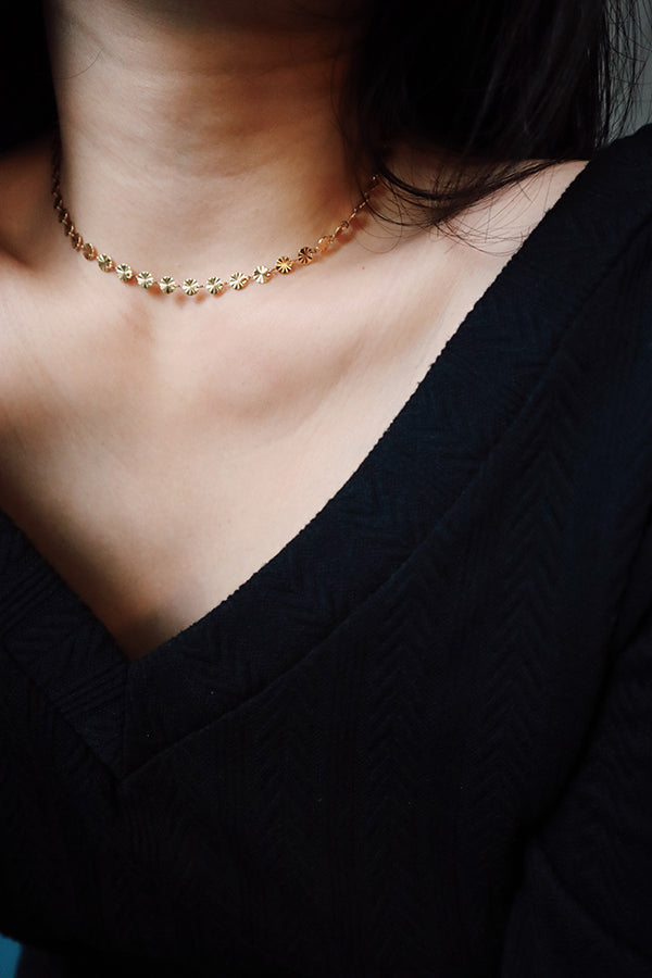 一位女性頸部和胸部的特寫,展示著Sienna Glow Choker——一條由小型、反光花朵狀鏈節製成的金色頸鍊,佩戴在她黑色上衣V領上方的頸部。