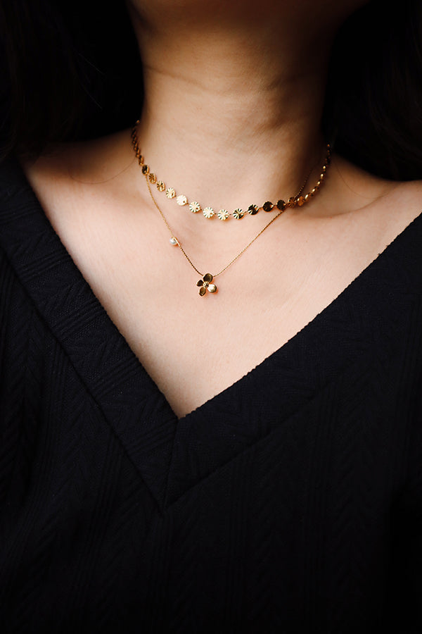 A close-up of a woman's neck, featuring the Sienna Glow Choker layered with a second gold necklace that has a small, four-petaled dark flower pendant. Both necklaces contrast sharply with the woman's black top.