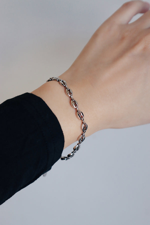 A close-up of a woman's forearm, showcasing a silver-toned bracelet with oval, coffee-bean links. The jewelry is worn against the edge of a black long-sleeved top.