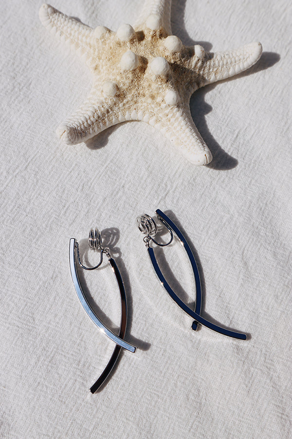 An overhead shot of a pair of silver-toned curved bar clip-on earrings, with a white starfish in the top background, resting on a white textured fabric. Sunlight casts shadows on the earrings and fabric.