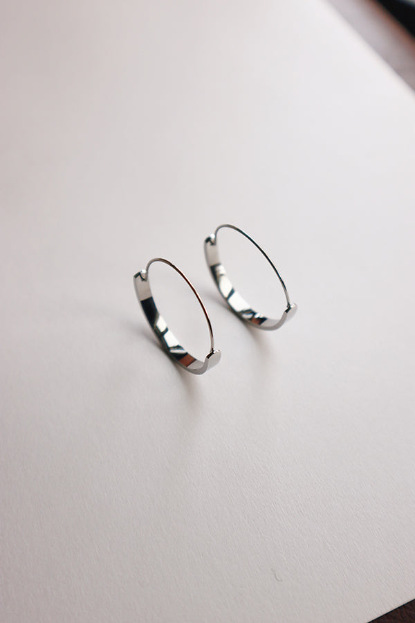An overhead shot of a pair of small, shiny silver hoop earrings with a flattened, open design, resting on a white surface. They cast subtle shadows.