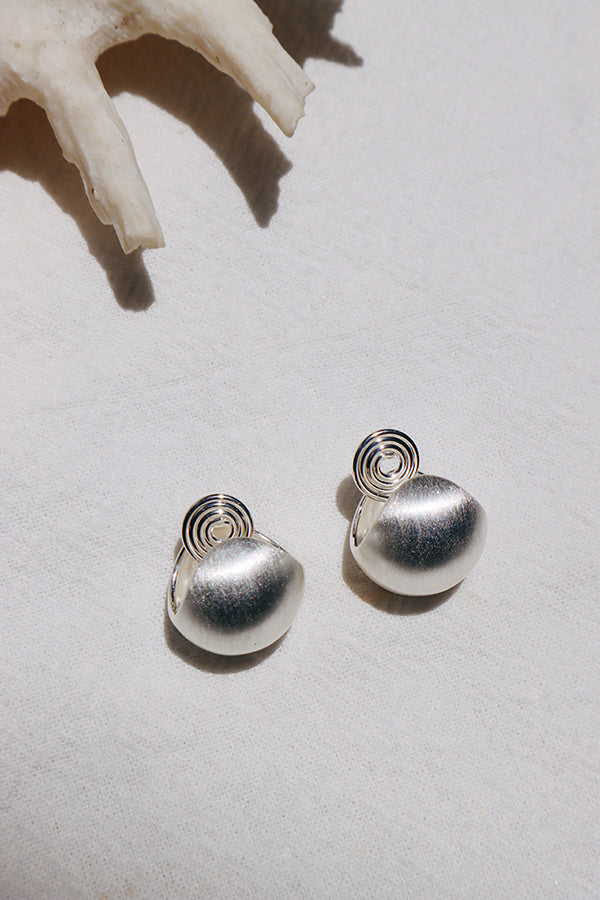 An overhead shot of a pair of silver-toned dome-shaped stud earrings with a brushed or textured finish, and a unique spiral backing visible. They are resting on a white textured fabric, with a large seashell partially visible in the top background.