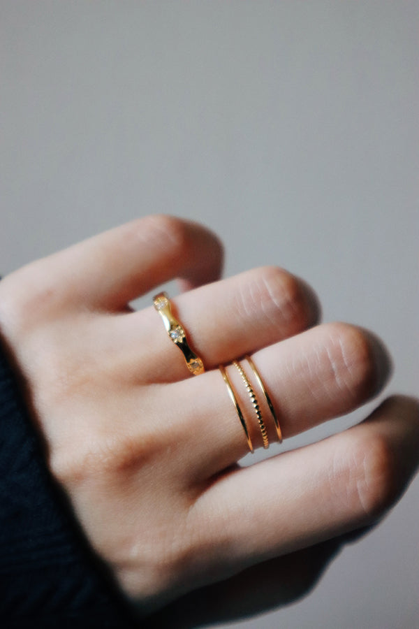 A very close-up, soft-focus shot of a woman's fingers, featuring the Liora Triple Band Ring on her middle finger and a gold wavy band with a central stone on her index finger. The woman is wearing a dark cuff.