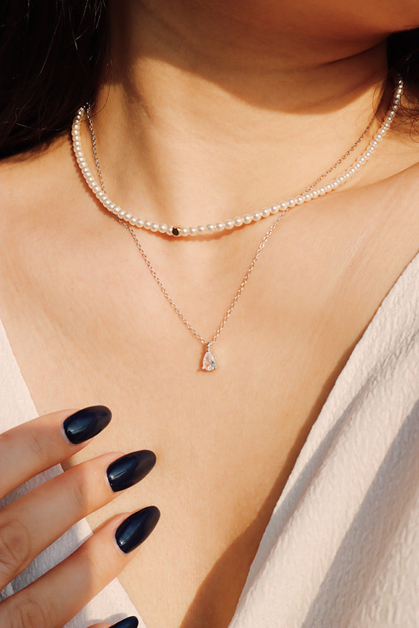 A close-up of a woman's chest and neck, wearing two layered necklaces. The top necklace is a string of small white pearls, and the bottom is a delicate silver chain with a pear-cut clear diamond or cubic zirconia pendant. Her hand, with dark blue manicured nails, is visible. The woman is wearing a white top.