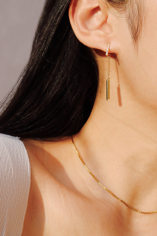 A close-up side profile of a woman's ear, showcasing a gold hoop earring with pave-set stones, from which dangles a thin gold bar on a delicate chain. The woman has long dark hair and is wearing a white ribbed top. A thin gold chain necklace is visible around her neck.