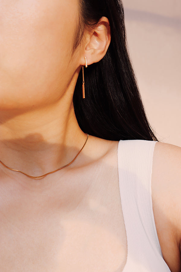 A close-up side profile of a woman's ear and shoulder, showcasing a gold hoop earring with pave-set stones, from which dangles a thin gold bar on a delicate chain. The woman has long dark hair and is wearing a white ribbed tank top. A thin gold chain necklace is also visible.