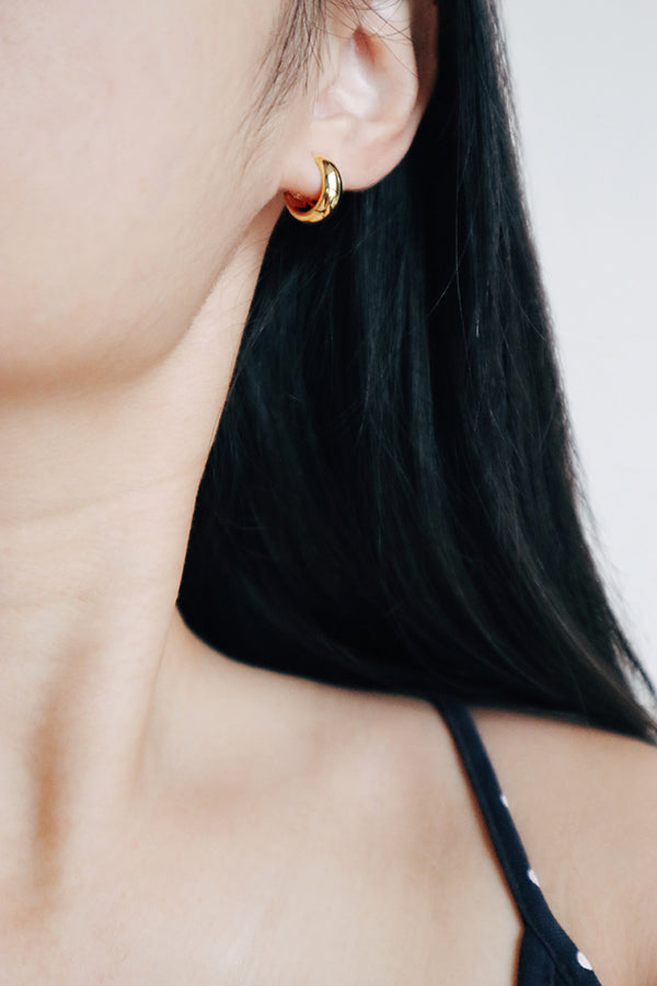 A close-up side profile of a woman's ear, showcasing a small, chunky, shiny gold hoop earring. The woman has long dark hair flowing around her ear and is wearing a dark top.