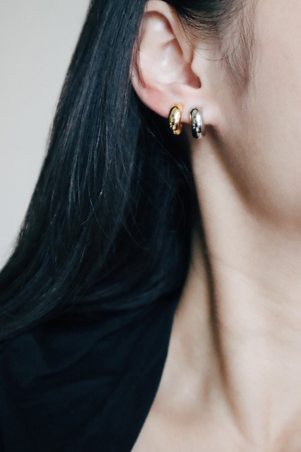 A close-up side profile of a woman's ear, showcasing a small gold chunky hoop earring in her left ear and a silver chunky hoop earring in her right ear (partially visible). The woman has long dark hair and is wearing a black top.