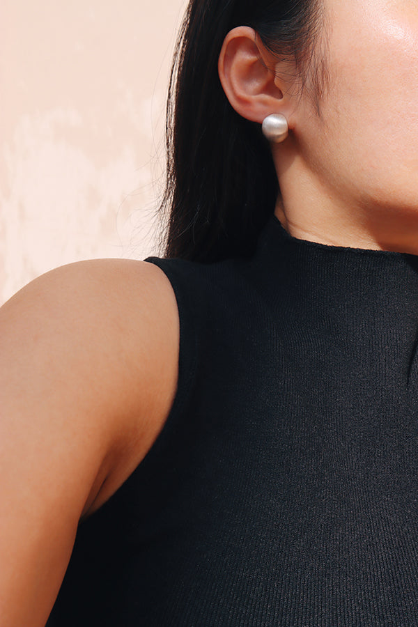 A close-up side profile of a woman's ear and shoulder, showcasing a large, shiny silver dome-shaped stud earring. The woman has long dark hair and is wearing a black ribbed sleeveless top.