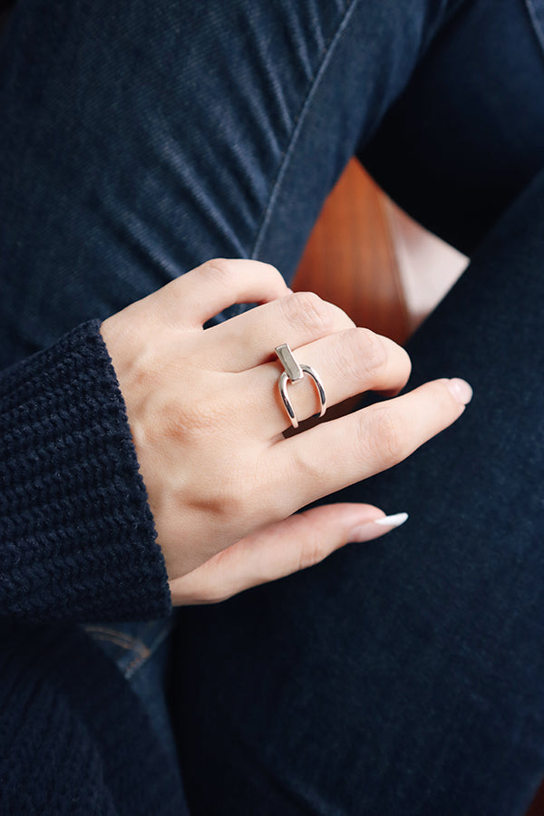 A close-up of a woman's hand, wearing a shiny silver ring with a unique bar and loop design on her ring finger. The woman is wearing a dark blue ribbed sweater sleeve and dark blue jeans.
