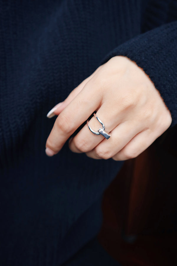 A close-up of a woman's hand, wearing a shiny silver ring with a unique bar and loop design on her ring finger. The woman has long, light-colored nails and is wearing a dark blue ribbed sweater. Her hand is resting on a dark wooden surface.
