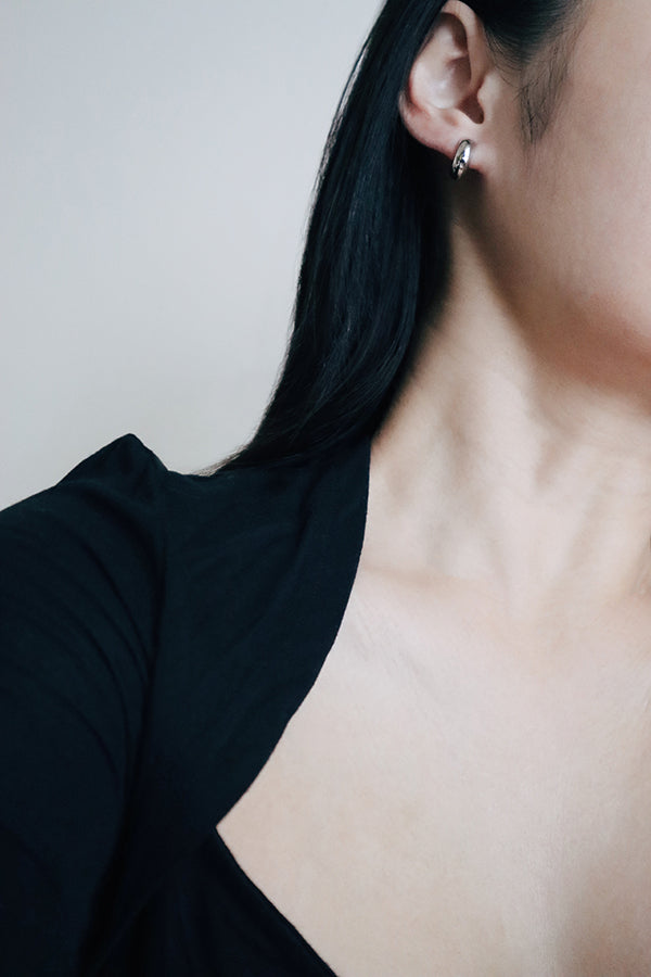 A close-up side profile of a woman's ear and shoulder, showcasing a small, chunky, shiny silver hoop earring. The woman has long dark hair flowing around her ear and is wearing a black blazer.
