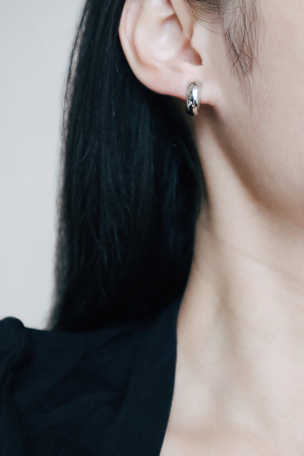 A close-up side profile of a woman's ear, showcasing a small, chunky, shiny silver hoop earring. The woman has long dark hair flowing around her ear and is wearing a dark top.