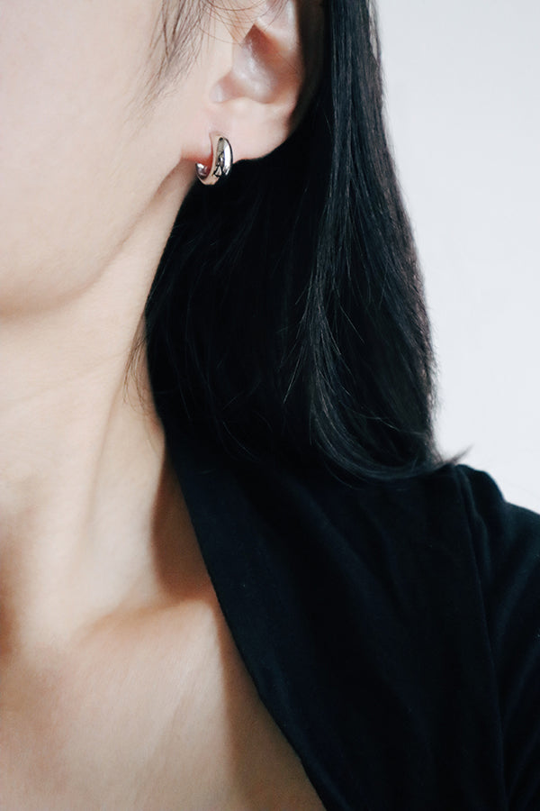 A close-up side profile of a woman's ear and shoulder, showcasing a small, chunky, shiny silver hoop earring. The woman has long dark hair flowing around her ear and is wearing a black blazer.