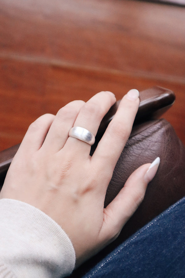 A close-up of a woman's hand with long, white French-tip manicured nails, wearing a wide, shiny silver dome ring on her ring finger. Her hand is resting on a brown leather armrest, with blue jeans visible in the background.