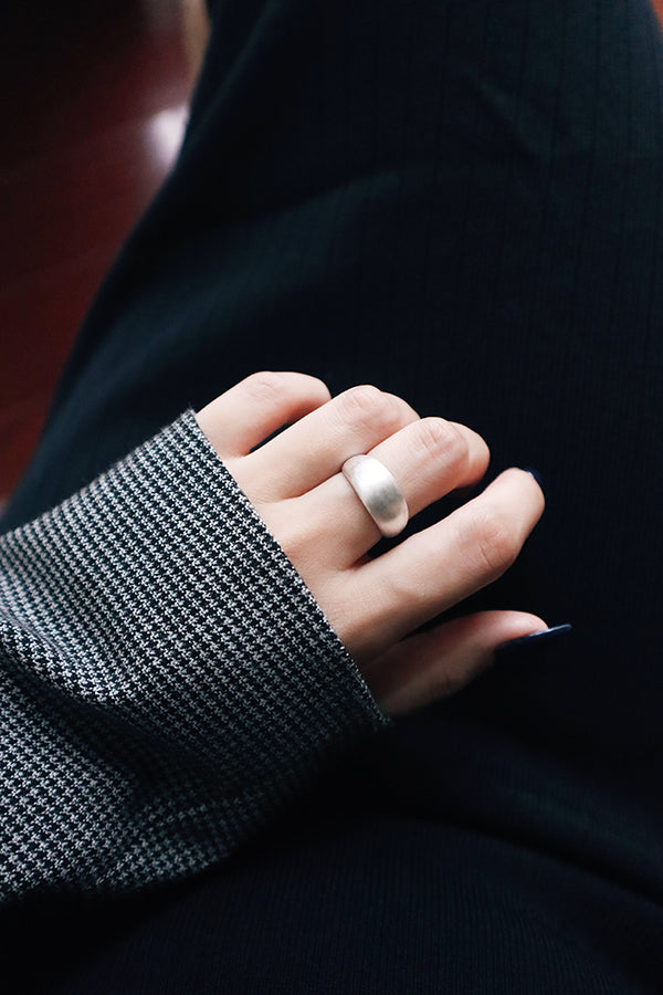 A close-up of a woman's hand with long, dark blue manicured nails, wearing a wide, shiny silver dome ring on her ring finger. The woman is wearing a black and white plaid blazer sleeve and a black garment.