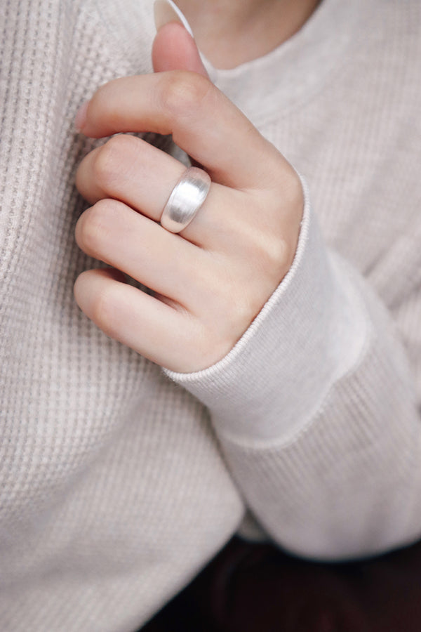 A close-up of a woman's hand, wearing a wide, shiny silver dome ring on her ring finger. Her hand is gently touching the cuff of her light cream-colored sweater.