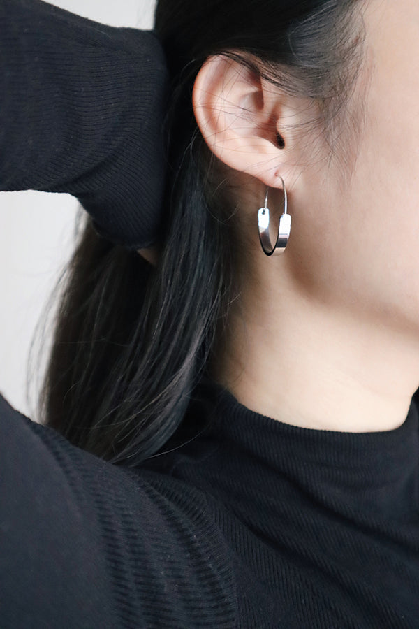 A close-up side profile of a woman's ear, showcasing a small, shiny silver hoop earring with a flattened, open design. Her hand is visible behind her ear, possibly adjusting her dark hair. The woman is wearing a black ribbed turtleneck.