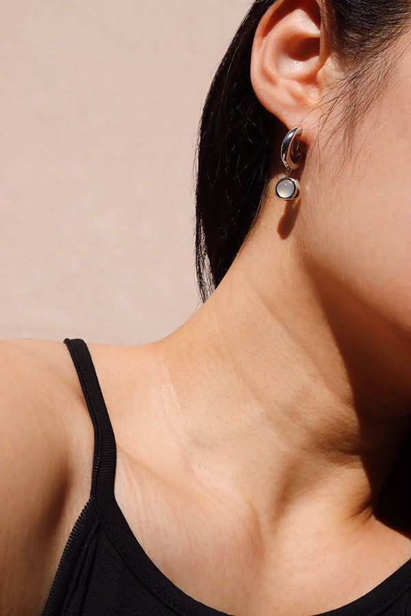 A close-up side profile of a woman's ear and neck, showcasing a shiny silver small hoop earring from which a round, iridescent white opalite bead dangles. The woman has long dark hair and is wearing a black tank top, with sunlight casting a distinct shadow on her neck.