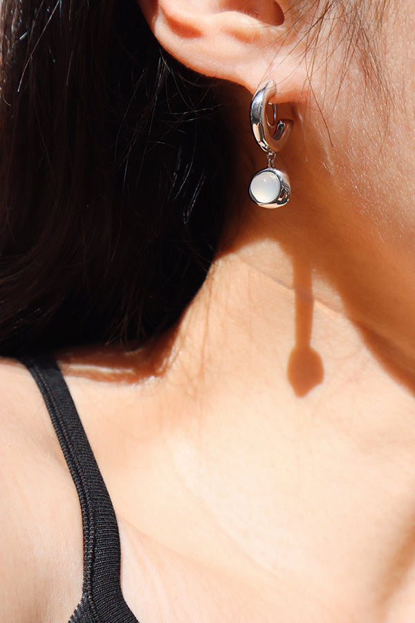 A close-up side profile of a woman's ear and shoulder, showcasing a shiny silver small hoop earring from which a round, iridescent white opalite bead dangles. The woman has long dark hair and is wearing a black tank top, with strong sunlight casting a long shadow from the earring onto her shoulder.