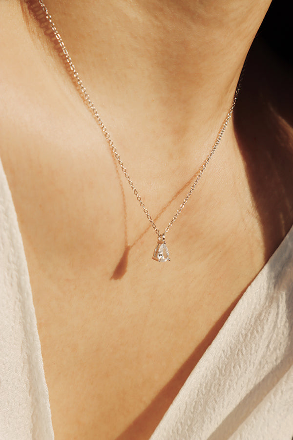 A close-up of a woman's chest and neck, wearing a delicate silver chain necklace with a single pear-cut clear diamond or cubic zirconia pendant. The woman is wearing a white top, with sunlight casting shadows.