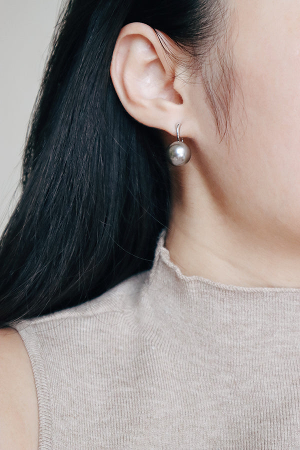 A close-up side profile of a woman's ear and neck, showcasing a silver-toned hoop earring with a dangling large silver-gray spherical pearl. The woman has long dark hair and is wearing a light beige ribbed turtleneck.