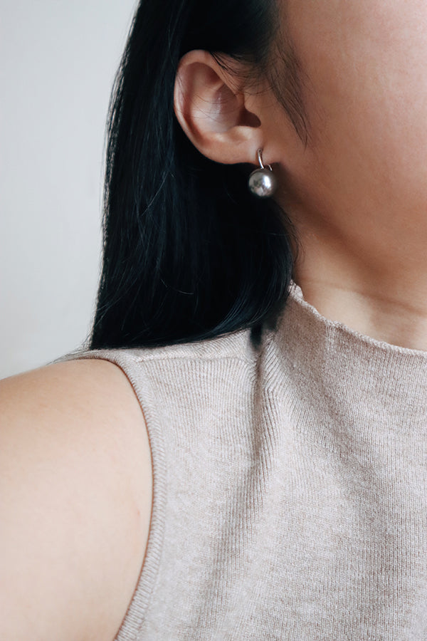A close-up side profile of a woman's ear and shoulder, showcasing a silver-toned hoop earring with a dangling large silver-gray spherical pearl. The woman has long dark hair and is wearing a light beige ribbed sleeveless top.
