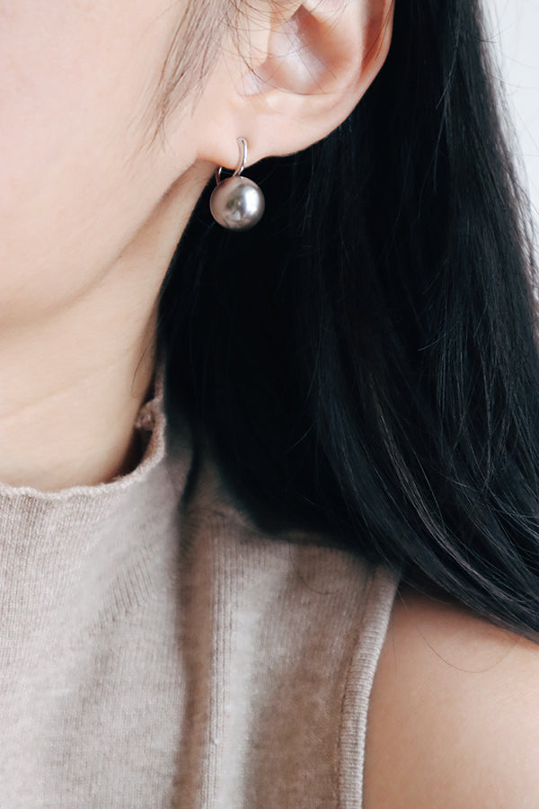 A close-up side profile of a woman's ear, showcasing a silver-toned hoop earring with a dangling large silver-gray spherical pearl. The woman has long dark hair and is wearing a light beige ribbed turtleneck.