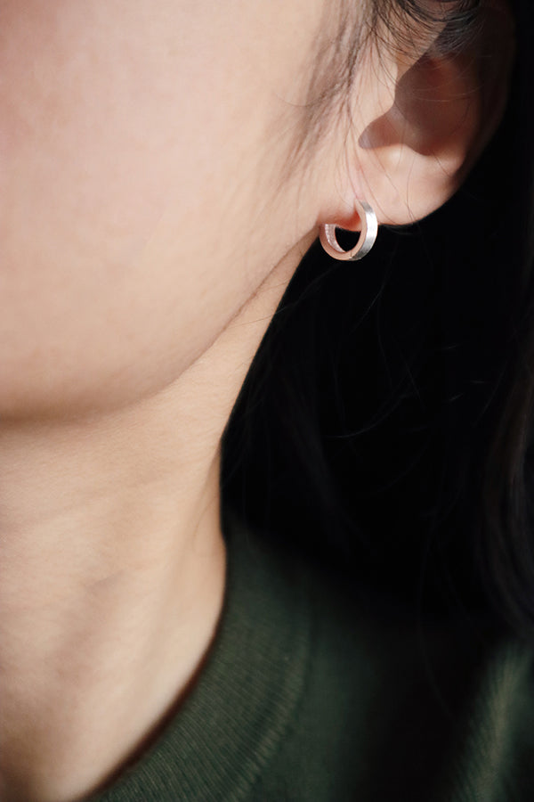A close-up side profile of a woman's ear, showcasing a small, shiny silver rounded hoop earring. The woman has long dark hair and is wearing a dark green top. Her neck is visible.