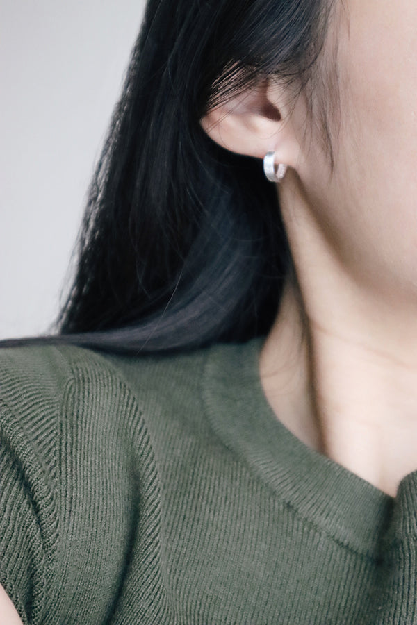 A close-up side profile of a woman's ear, showcasing a small, shiny silver rounded hoop earring. The woman has long dark hair and is wearing a dark green ribbed top.
