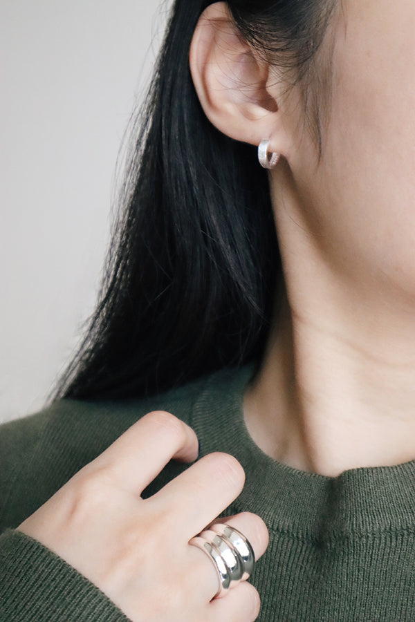 A close-up side profile of a woman's ear and hand, showcasing a small, shiny silver rounded hoop earring. On her ring finger, she is wearing a chunky layered silver ring. The woman has long dark hair and is wearing a dark green ribbed sweater.
