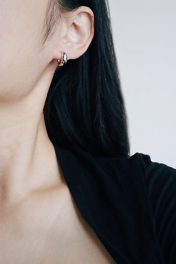 A close-up side profile of a woman's ear, showcasing a small, chunky, shiny silver hoop earring. The woman has long dark hair flowing around her ear and is wearing a black top.