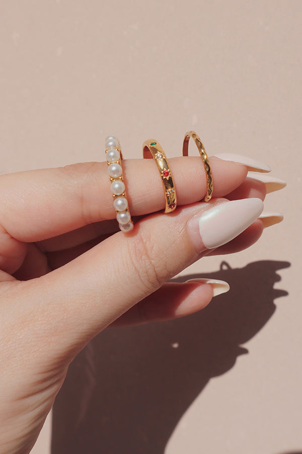 A hand wearing three dainty gold rings stacked on one finger: a full pearl band ring, a gold band with colorful cubic zirconia stones, and a delicate twisted gold ring.