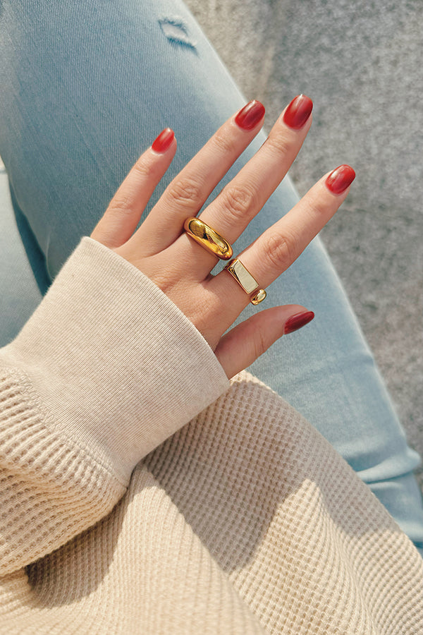 A hand wearing two chunky, bold gold statement rings: a smooth dome ring and an open-ended geometric ring with a rectangular bar and a sphere.