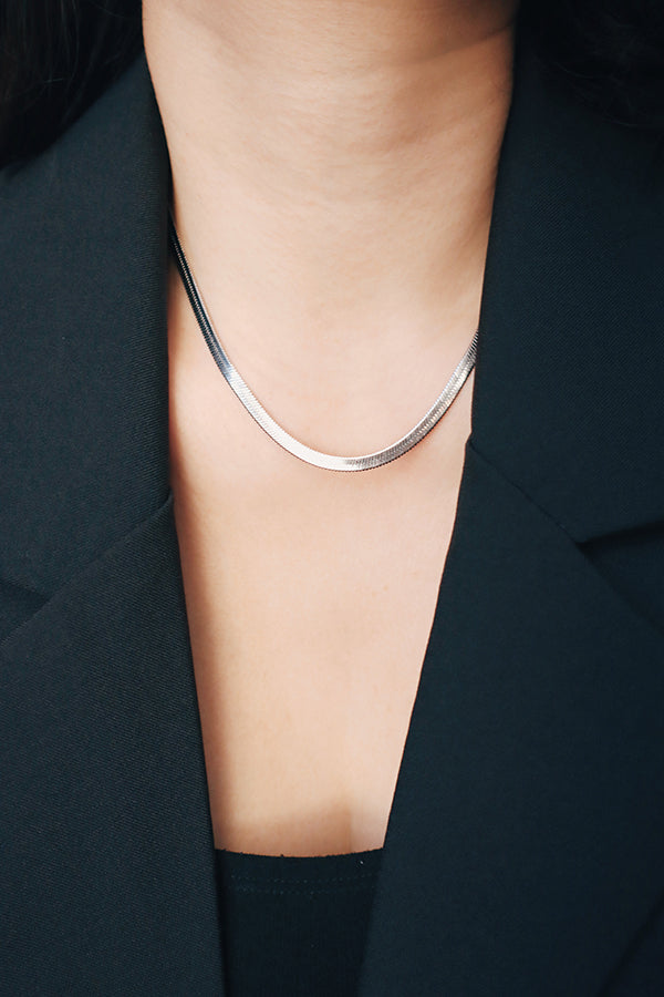 A close-up of a woman's neck and chest, wearing a flat, shiny silver herringbone chain necklace. The woman is wearing a black blazer.