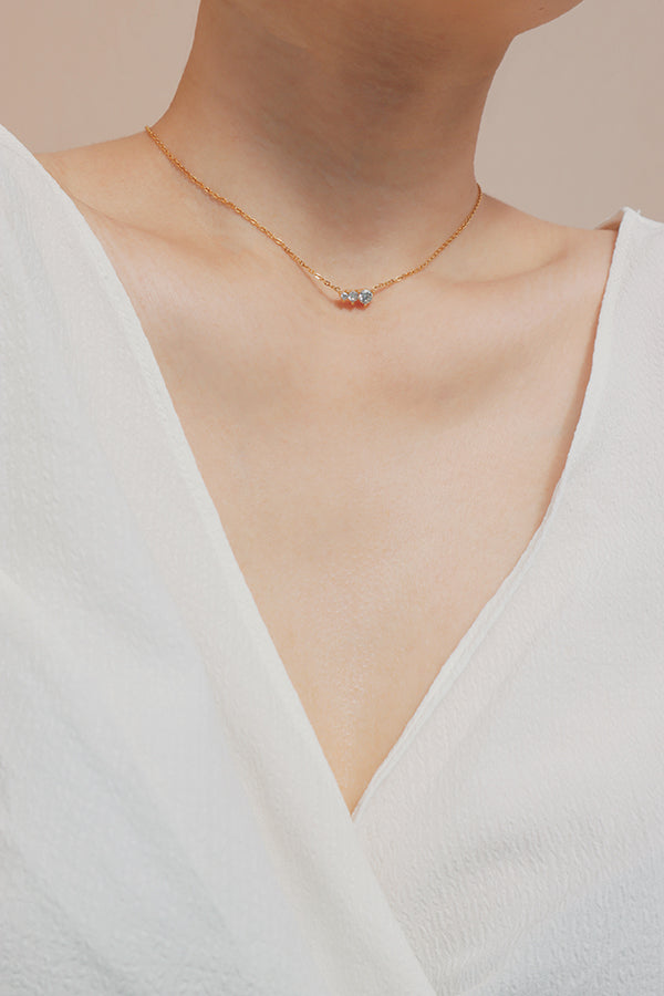 A delicate gold necklace with a horizontal cluster of three clear cubic zirconia stones, worn on a person's neck over a white v-neck top.