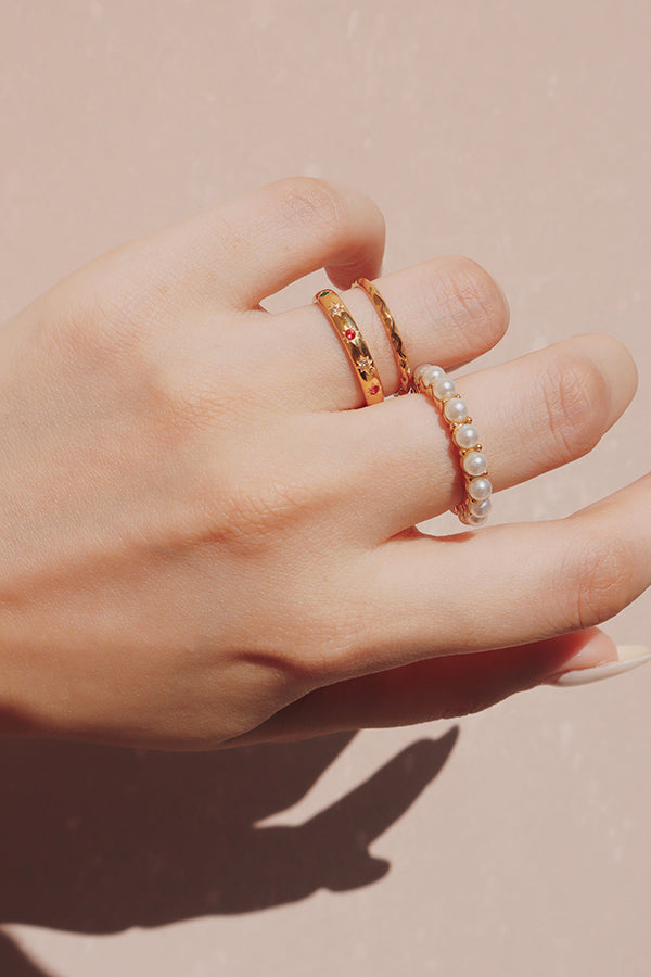 一隻手特寫,疊戴三枚金色戒指:一枚珍珠鑲嵌戒指、一枚鑲有多彩鋯石的金色戒指,以及一枚精緻扭紋金戒。