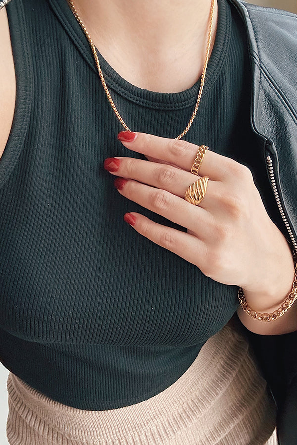 A woman's hand wearing two gold rings: a delicate chain ring and a bold, ribbed croissant-style ring, styled with a gold chain necklace and bracelet.