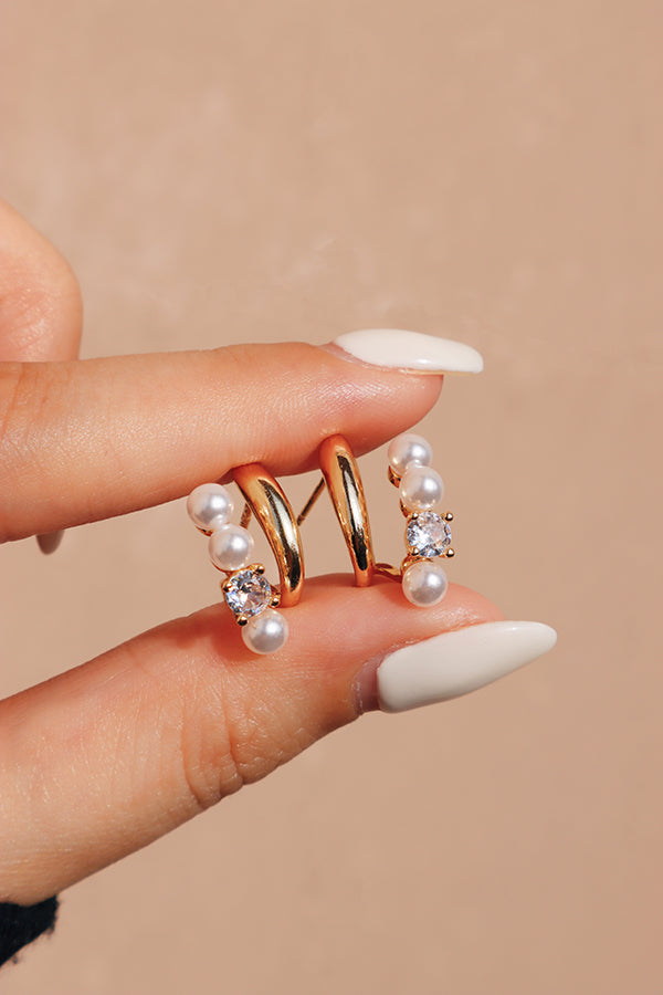 A delicate pair of gold hoop earrings with pearls and sparkling zircon accents, held between fingers against a neutral background.