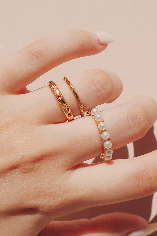 Close-up of a hand wearing three gold rings: a pearl-studded band, a ring featuring red gemstones, and a twisted gold band, creating a chic stacked look.