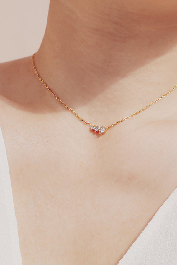 一條簡約經典的精緻金色項鍊,水平鑲嵌著三顆透明鋯石,佩戴於頸部。